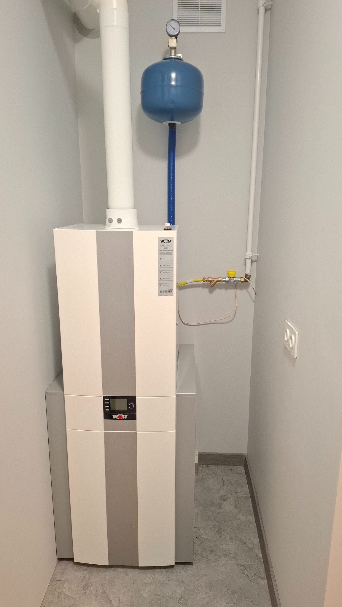 Kompaktowy, biało-szary piec gazowy marki Wolf z niebieskim naczyniem przeponowym w małej kotłowni z szarą podłogą i białymi rurami. Widoczny zawór gazowy i gniazdko elektryczne.