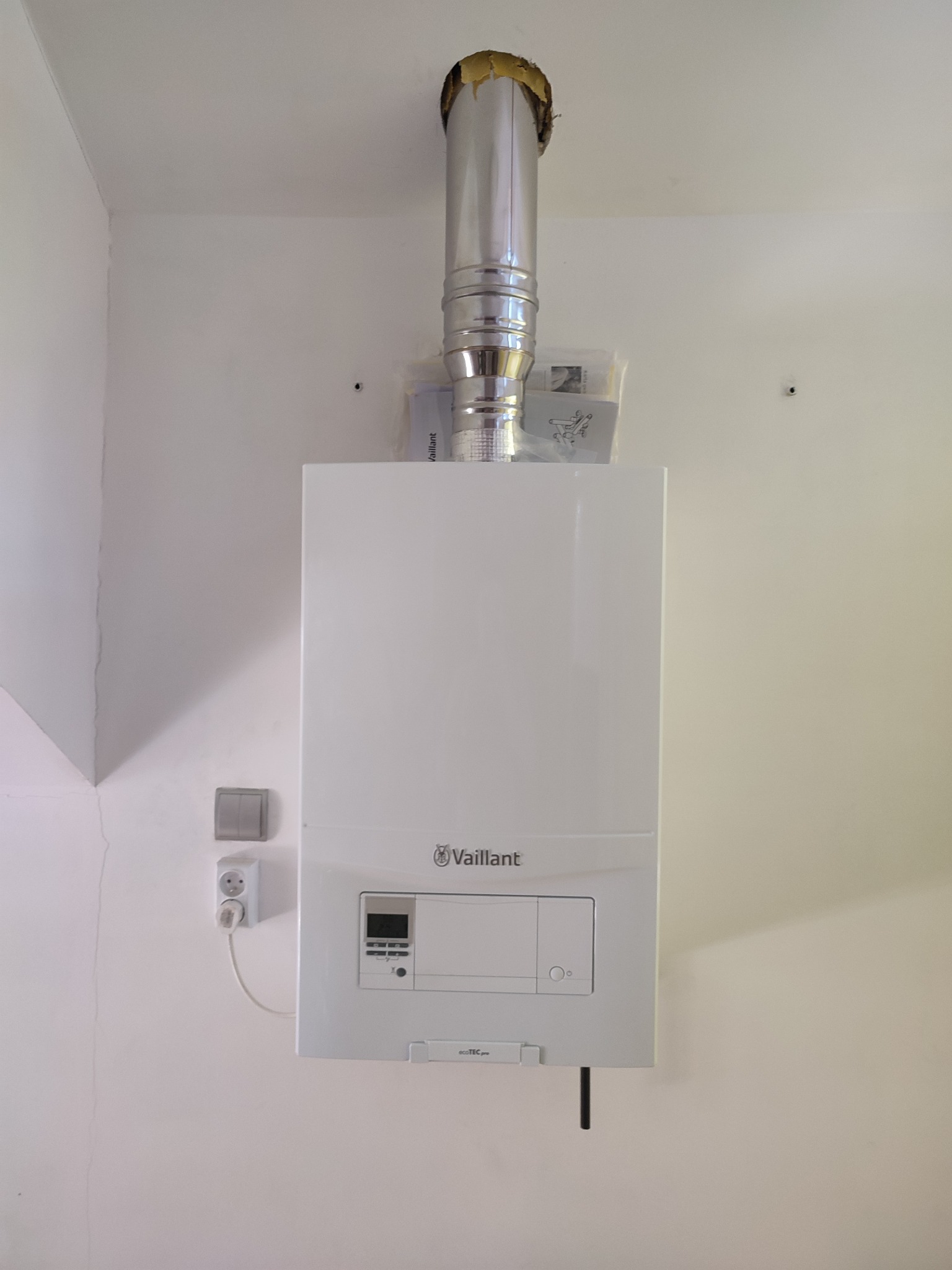 Biały, wiszący piec gazowy Vaillant z kominem, świeżo zamontowany na białej ścianie w Gdyni. Widoczne gniazdko elektryczne i instrukcja obsługi.