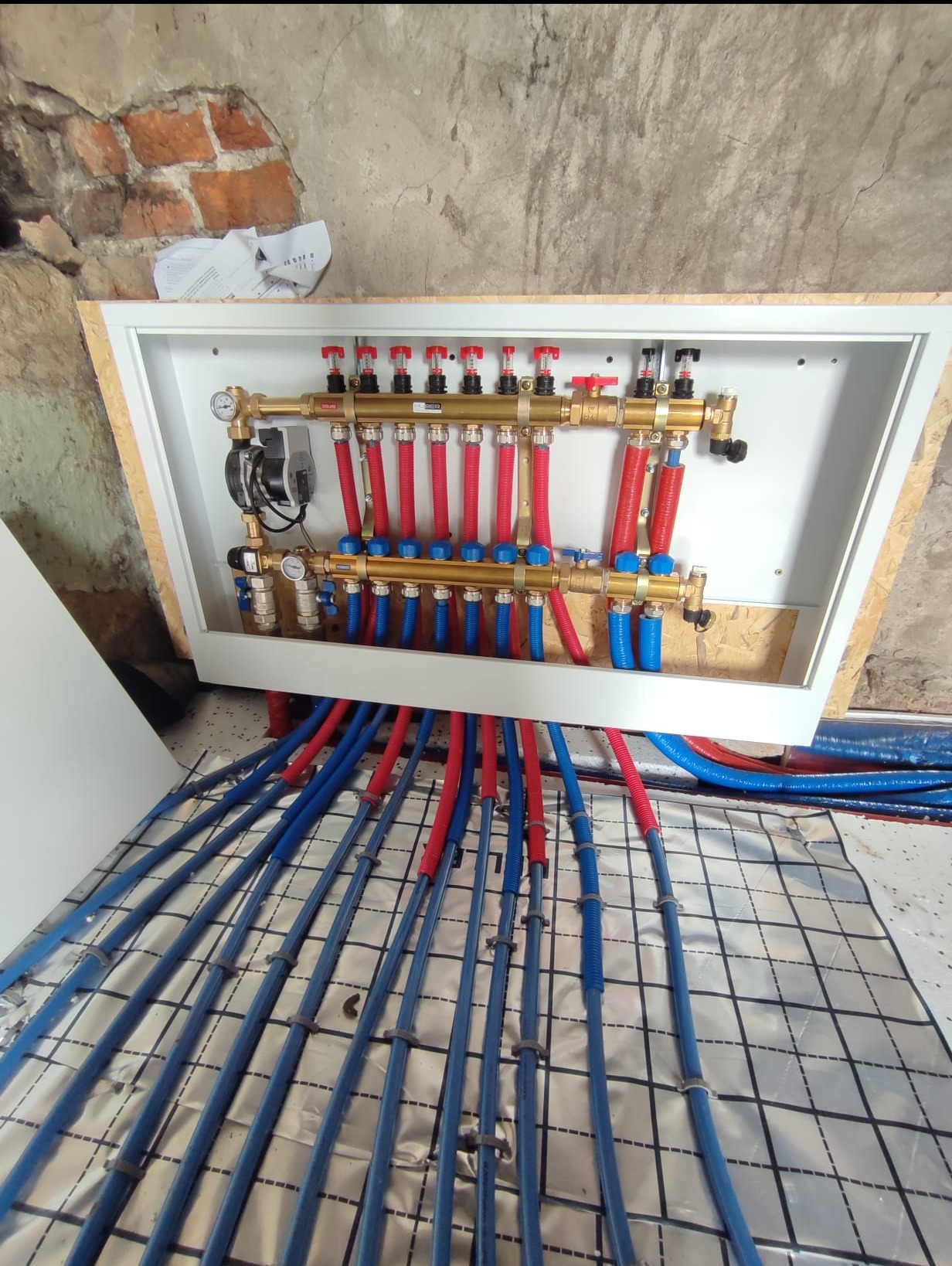 Rozdzielacz ogrzewania podłogowego z rurami PEX w kolorze niebieskim i czerwonym, zamontowany w białej skrzynce. Widoczne elementy instalacji i folia termoizolacyjna.
