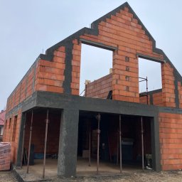 SKYRISE SPÓŁKA Z OGRANICZONĄ ODPOWIEDZIALNOŚCIĄ - Surowy stan domu murowanego z pustaków ceramicznych, widoczne stalowe podpory podtrzymujące strop, zapakowane pustaki obok budynku, szary dzień.