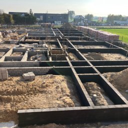 SKYRISE SPÓŁKA Z OGRANICZONĄ ODPOWIEDZIALNOŚCIĄ - Szeroki kadr na plac budowy z uformowanymi fundamentami pod dom jednorodzinny, widoczne zbrojenia i częściowo wymurowane ściany fundamentowe z bloczków betonowych, w tle budynki i zieleń.