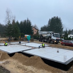 SKYRISE SPÓŁKA Z OGRANICZONĄ ODPOWIEDZIALNOŚCIĄ - Przygotowanie szalunku i zbrojenia płyty fundamentowej pod budowę domu jednorodzinnego, widoczna betoniarka i pracownicy w odblaskowych kamizelkach, pochmurny dzień.