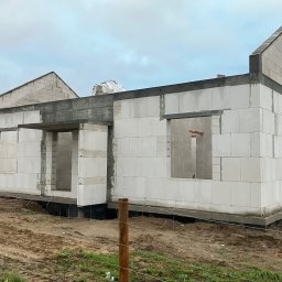 SKYRISE SPÓŁKA Z OGRANICZONĄ ODPOWIEDZIALNOŚCIĄ - Surowy stan domu z betonu komórkowego w trakcie budowy, widoczne otwory okienne i drzwiowe, teren wokół w stanie naturalnym, pochmurne niebo.