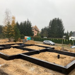 SKYRISE SPÓŁKA Z OGRANICZONĄ ODPOWIEDZIALNOŚCIĄ - Zalane betonem szalunki fundamentowe z wystającymi prętami zbrojeniowymi, wypełnione piaskiem na działce budowlanej z widocznym samochodem osobowym i przyczepą toaletową w tle.