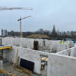 SKYRISE SPÓŁKA Z OGRANICZONĄ ODPOWIEDZIALNOŚCIĄ - Wnętrze powstającego budynku z białych bloczków, widoczne dwie osoby w odblaskowych kamizelkach i żółtym kasku, w tle żurawie budowlane i zabudowa miejska.
