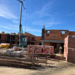 Budowa domu murowanego w trakcie wznoszenia ścian, widoczne palety z cegłami CERPOL, zbrojenia, pracownicy w odblaskowych kamizelkach i żuraw na tle błękitnego nieba.