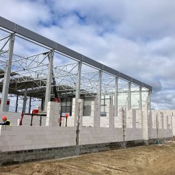 SKYRISE SPÓŁKA Z OGRANICZONĄ ODPOWIEDZIALNOŚCIĄ - Wznoszenie ścian z białych bloczków na budowie hali przemysłowej z widoczną stalową konstrukcją szkieletową i pracownikami w odblaskowych kamizelkach.