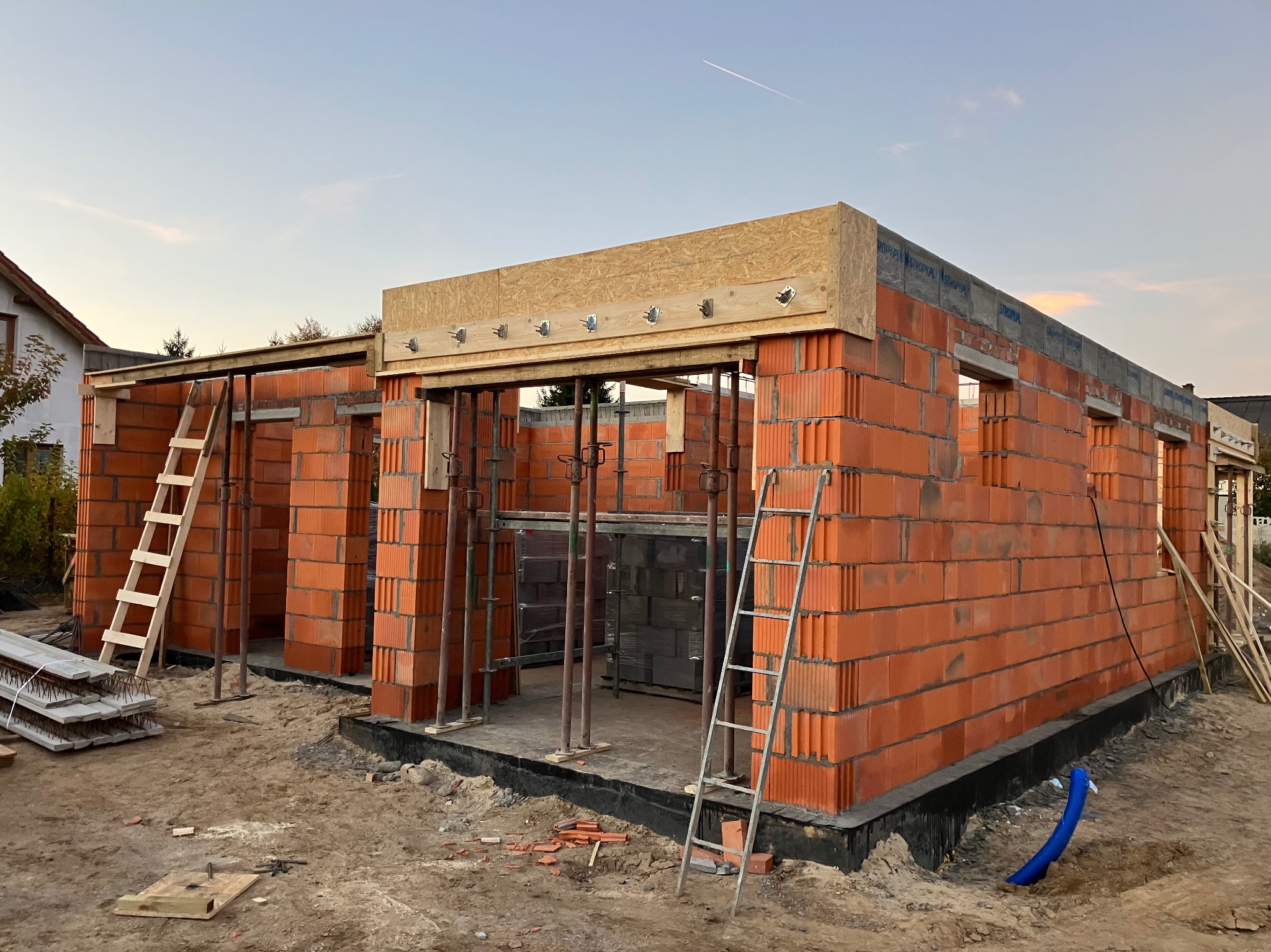 Budowa domu w trakcie wznoszenia ścian z czerwonej cegły, widoczne drewniane szalunki, podpory i drabiny, na ziemi gruz i fragment niebieskiej rury. W tle niebo z widocznym śladem po samolocie.