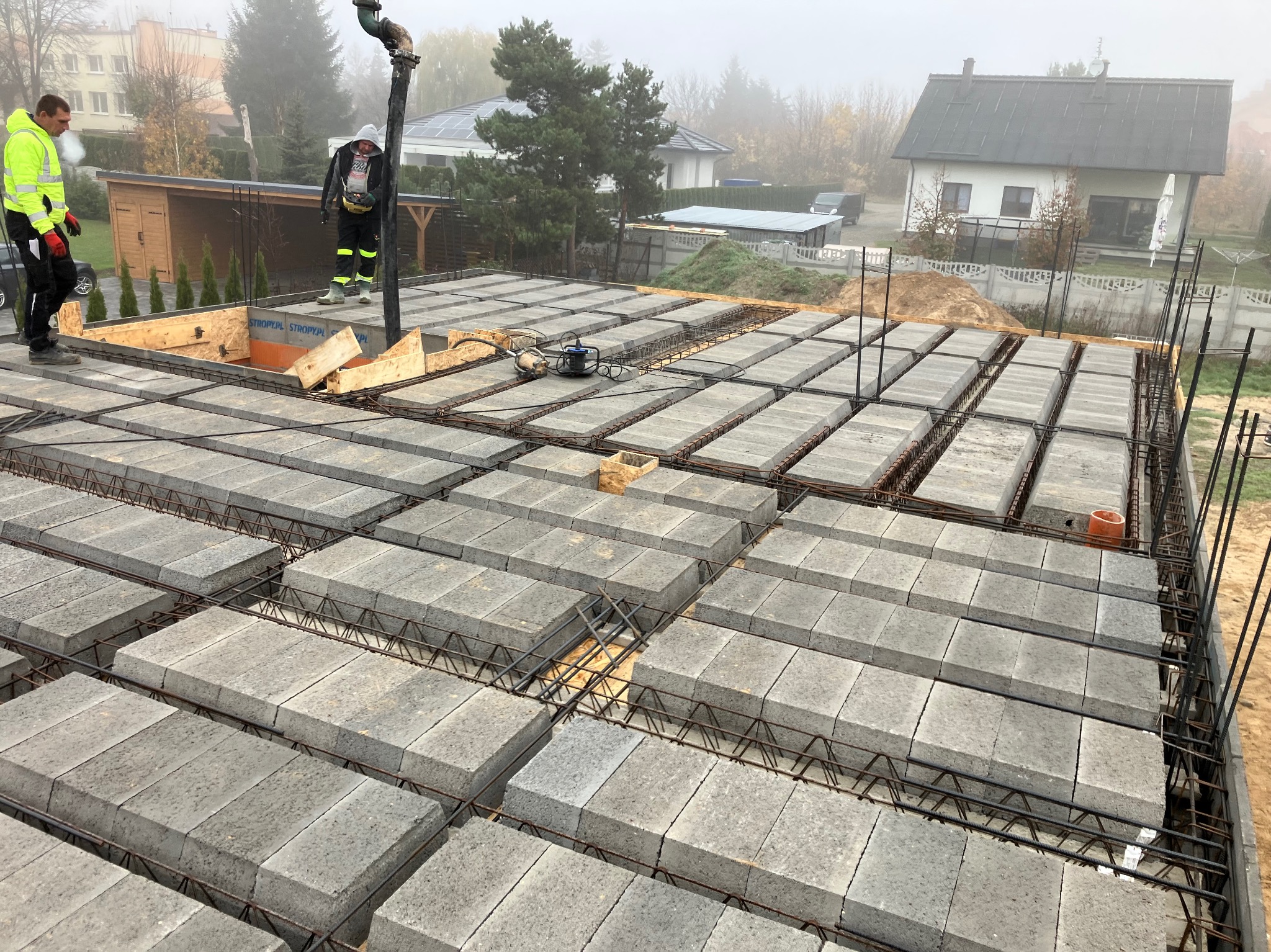 Budowa stropu Teriva: widok z góry na ułożone pustaki stropowe i zbrojenie, dwóch pracowników w odblaskowych ubraniach obsługuje pompę do betonu, w tle zabudowa jednorodzinna w porannej mgle.