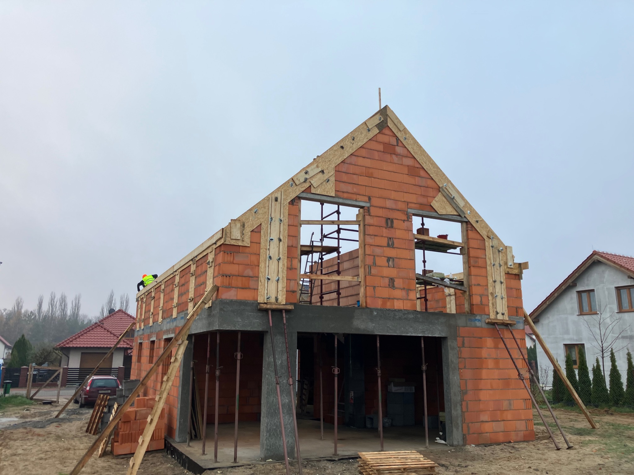 Wznoszenie dwukondygnacyjnego domu z czerwonej cegły kratówki; widoczne szalunki okienne i drewniane elementy konstrukcyjne dachu; pracownik w kamizelce odblaskowej na górze ściany.