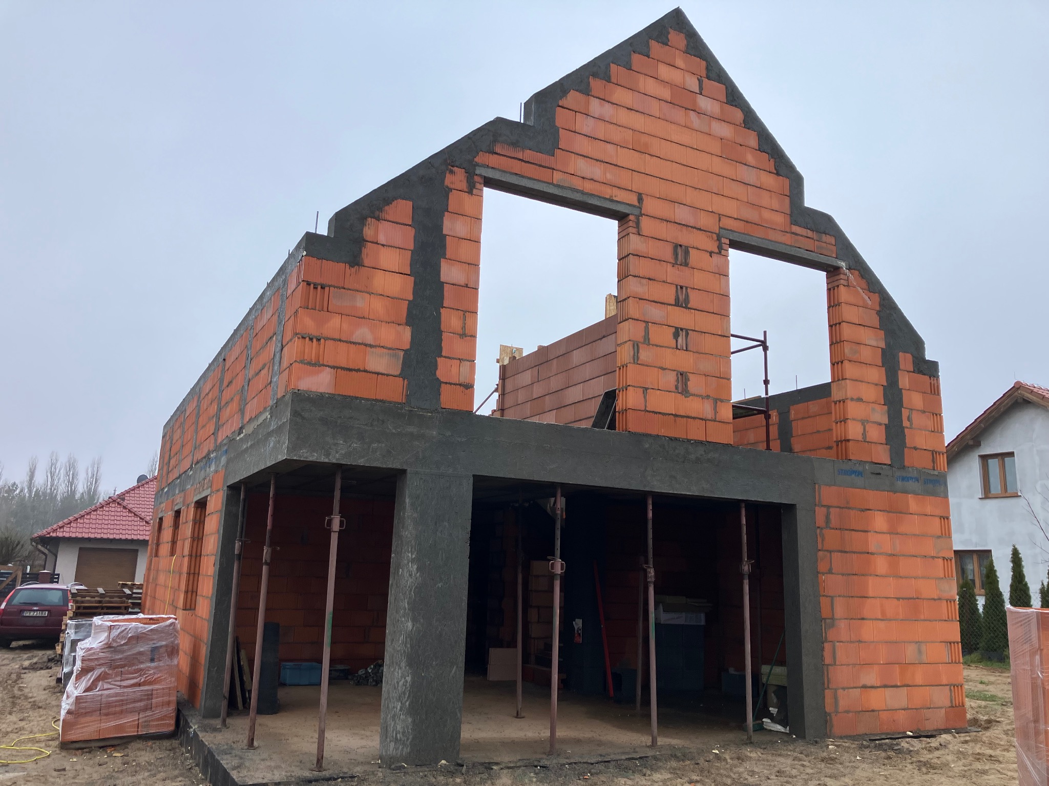 Surowy stan domu murowanego z pustaków ceramicznych, widoczne stalowe podpory podtrzymujące strop, zapakowane pustaki obok budynku, szary dzień.