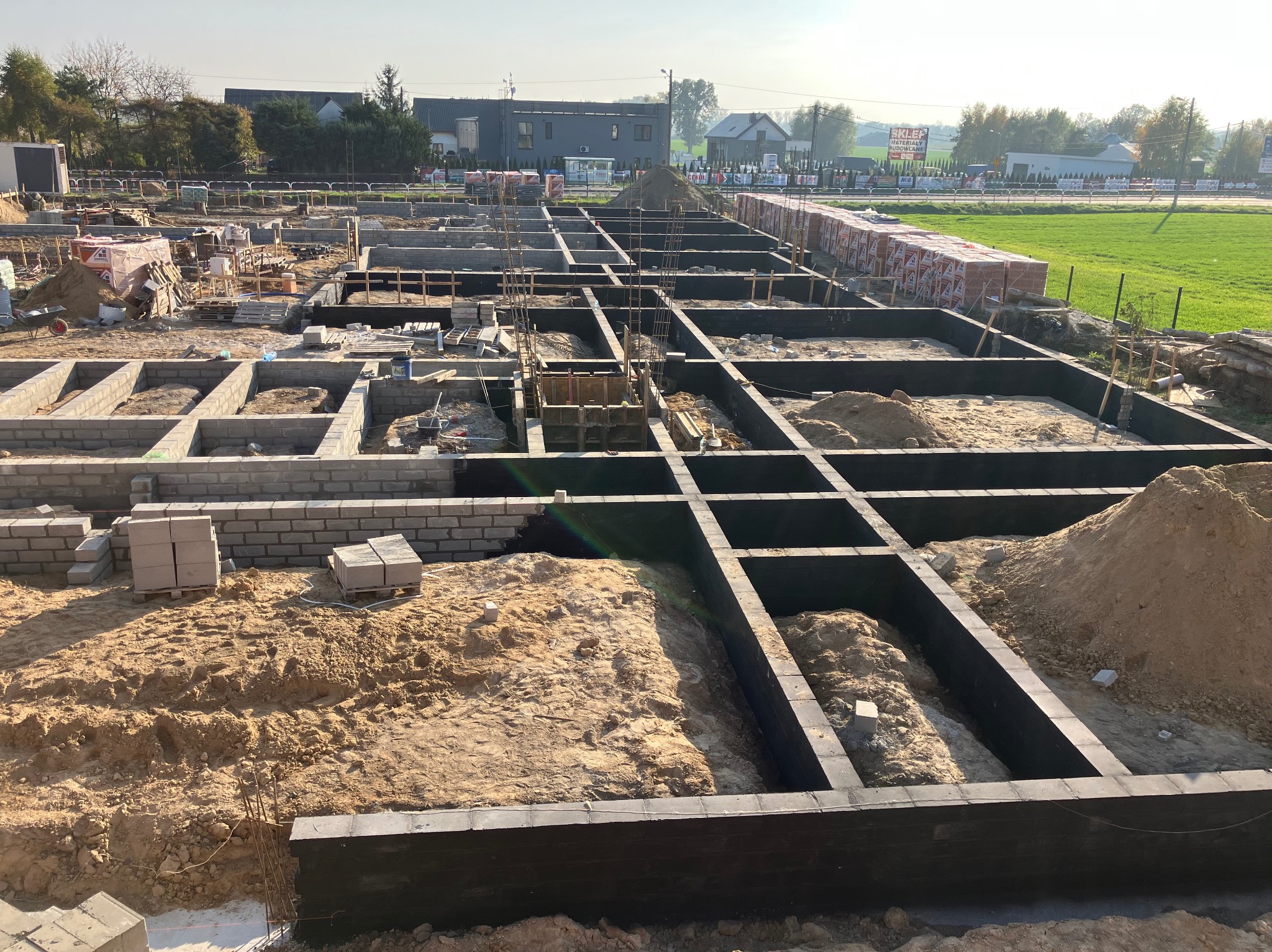 Szeroki kadr na plac budowy z uformowanymi fundamentami pod dom jednorodzinny, widoczne zbrojenia i częściowo wymurowane ściany fundamentowe z bloczków betonowych, w tle budynki i zieleń.
