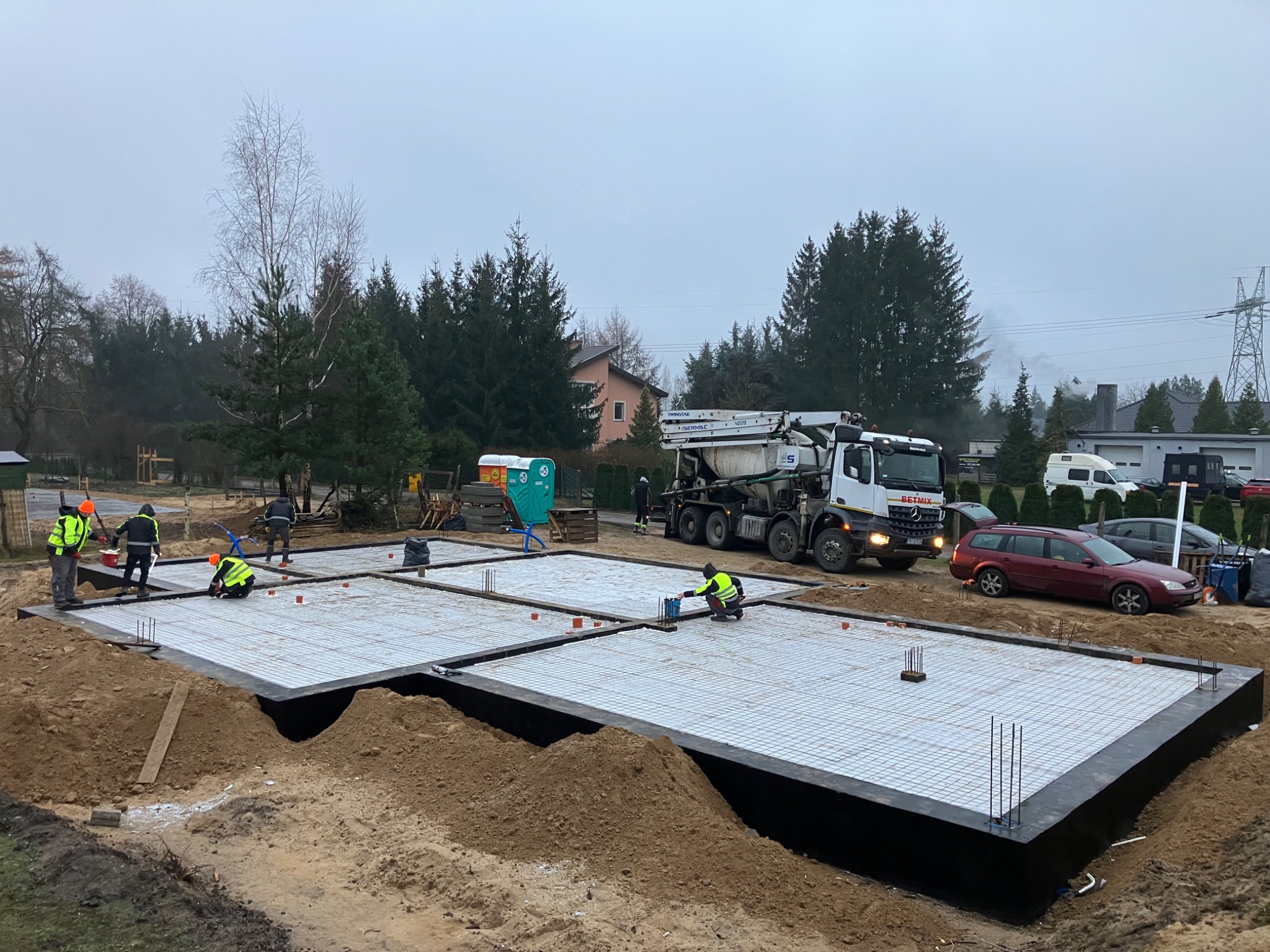 Przygotowanie szalunku i zbrojenia płyty fundamentowej pod budowę domu jednorodzinnego, widoczna betoniarka i pracownicy w odblaskowych kamizelkach, pochmurny dzień.