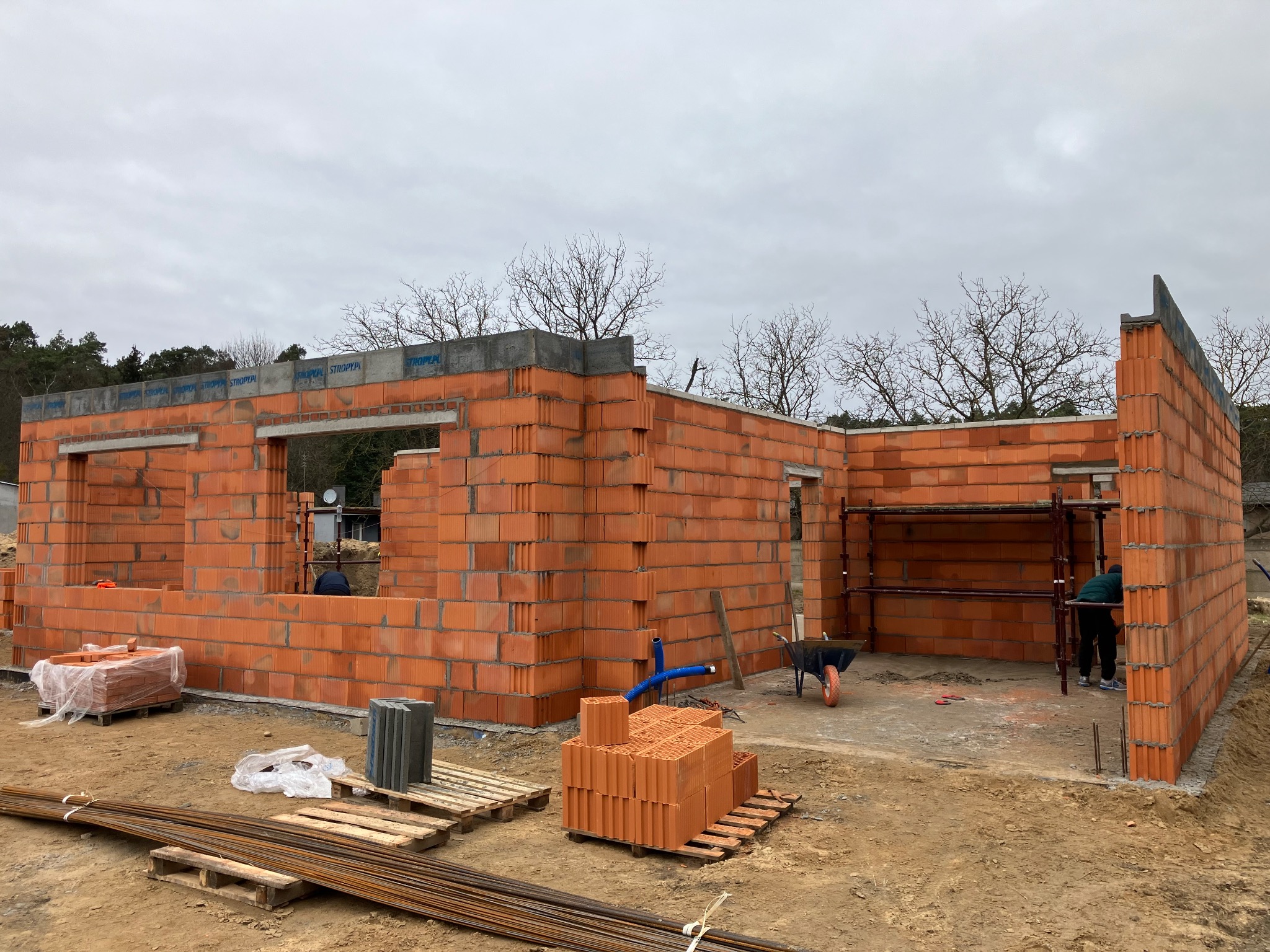 Wznoszenie ścian domu z czerwonej cegły ceramicznej, widoczne rusztowania, palety z cegłami oraz pracownik wewnątrz budynku w trakcie prac murarskich.