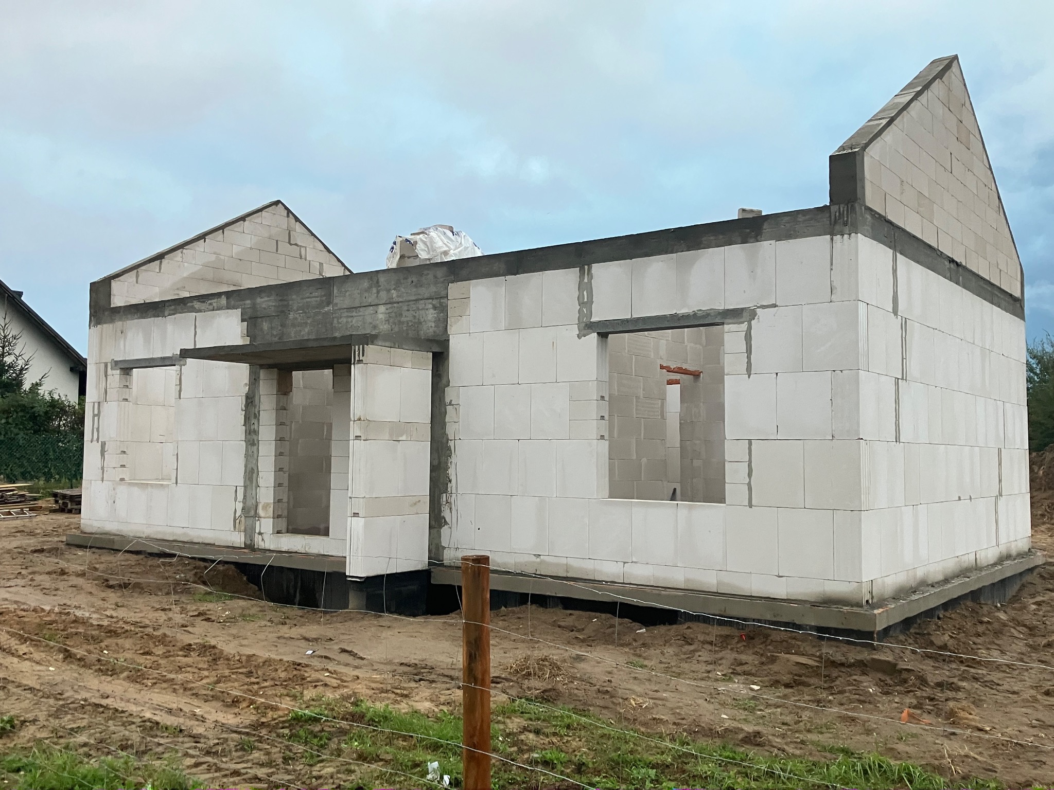 Surowy stan domu z betonu komórkowego w trakcie budowy, widoczne otwory okienne i drzwiowe, teren wokół w stanie naturalnym, pochmurne niebo.