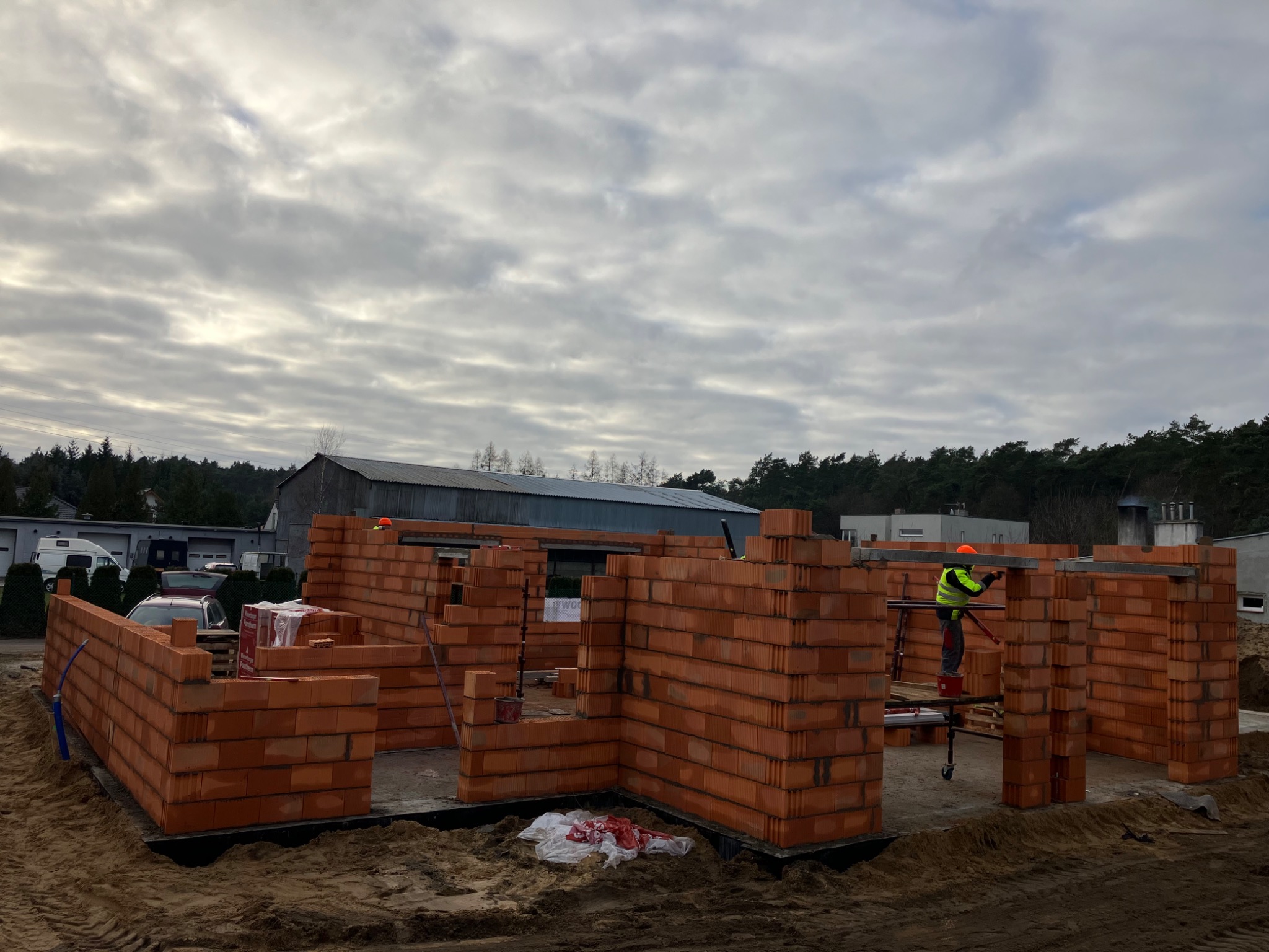 Budowa domu z czerwonej cegły Porotherm, widoczny fragment ściany z otworami okiennymi, pracownik w żółtej kamizelce na rusztowaniu, pochmurne niebo w tle.