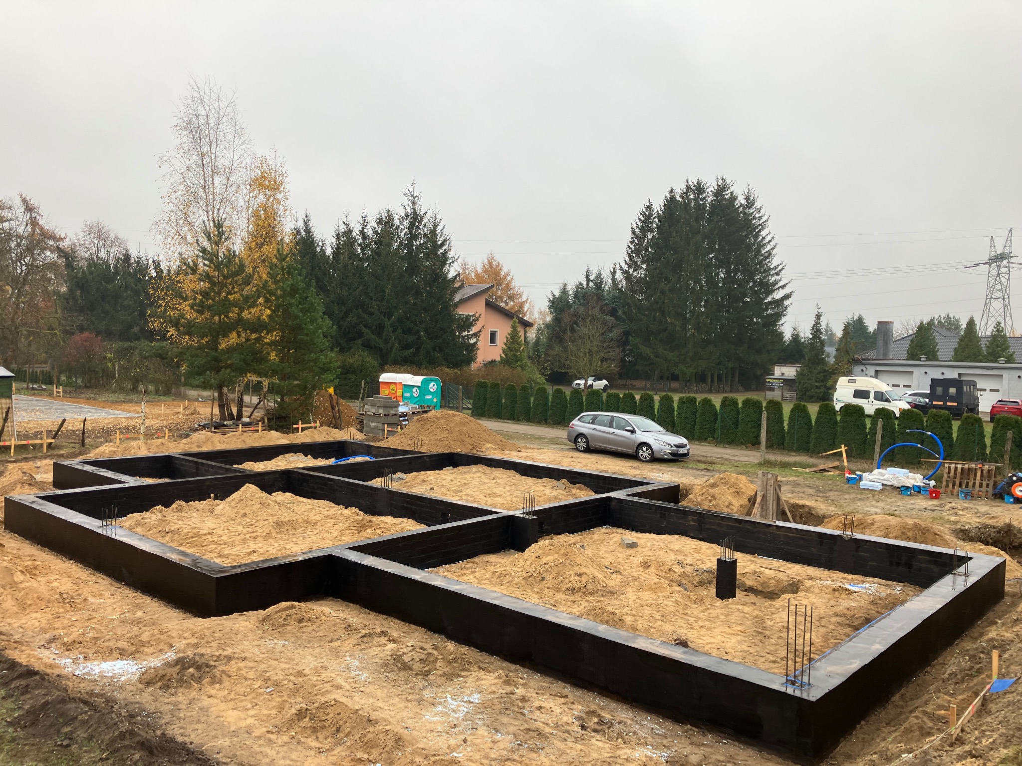 Zalane betonem szalunki fundamentowe z wystającymi prętami zbrojeniowymi, wypełnione piaskiem na działce budowlanej z widocznym samochodem osobowym i przyczepą toaletową w tle.