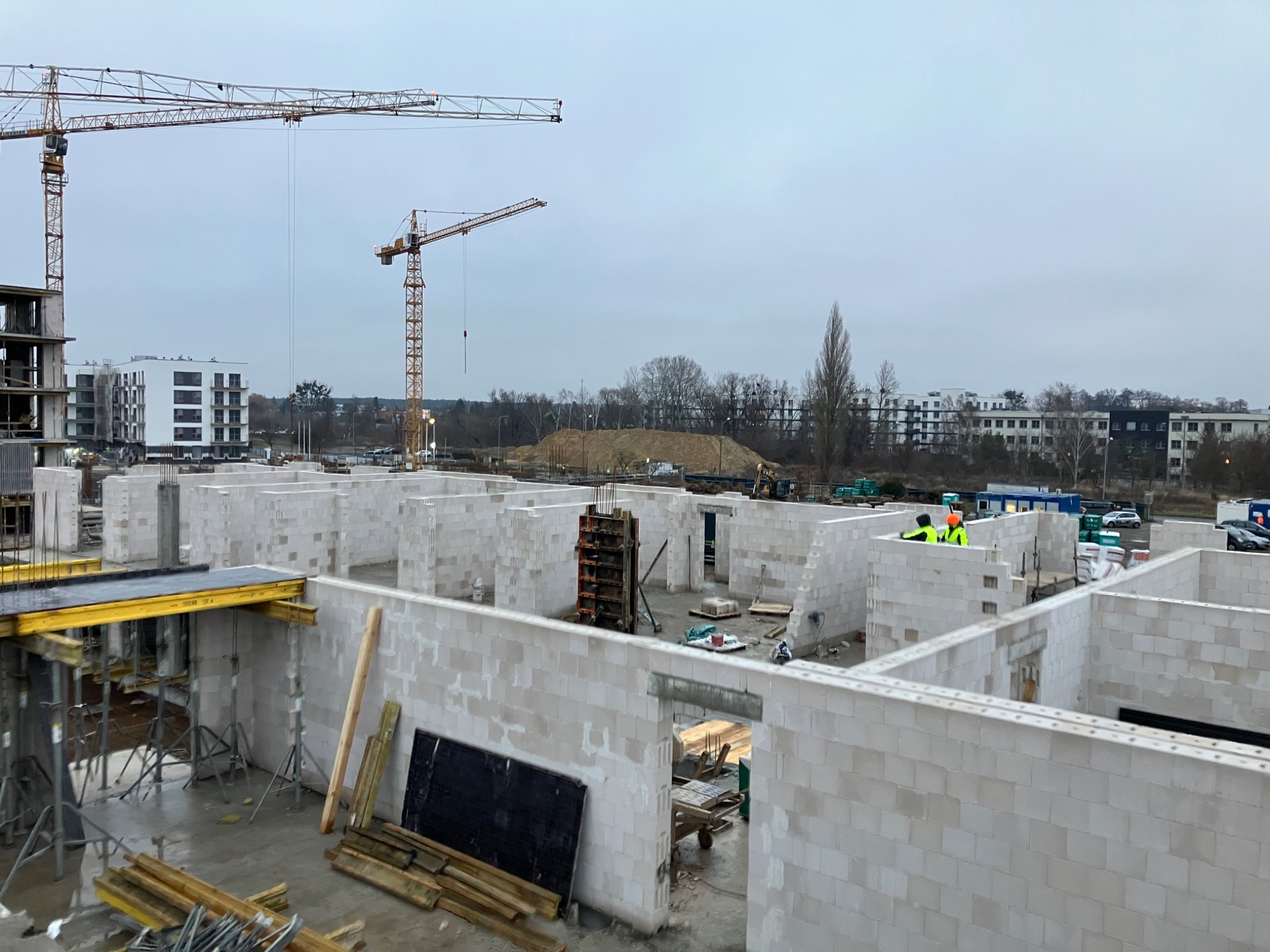 Wnętrze powstającego budynku z białych bloczków, widoczne dwie osoby w odblaskowych kamizelkach i żółtym kasku, w tle żurawie budowlane i zabudowa miejska.