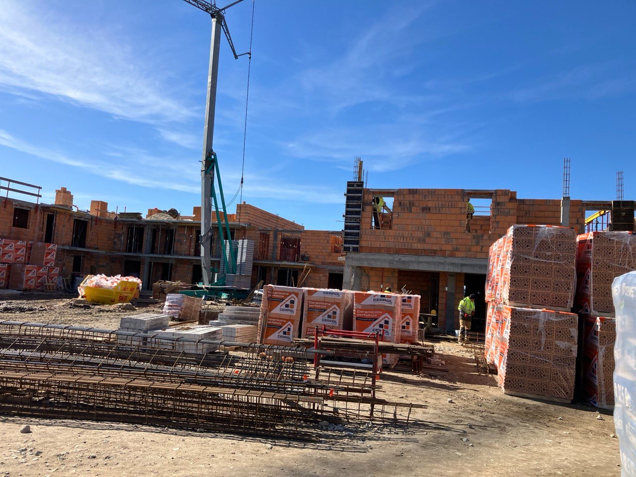 Budowa domu murowanego w trakcie wznoszenia ścian, widoczne palety z cegłami CERPOL, zbrojenia, pracownicy w odblaskowych kamizelkach i żuraw na tle błękitnego nieba.