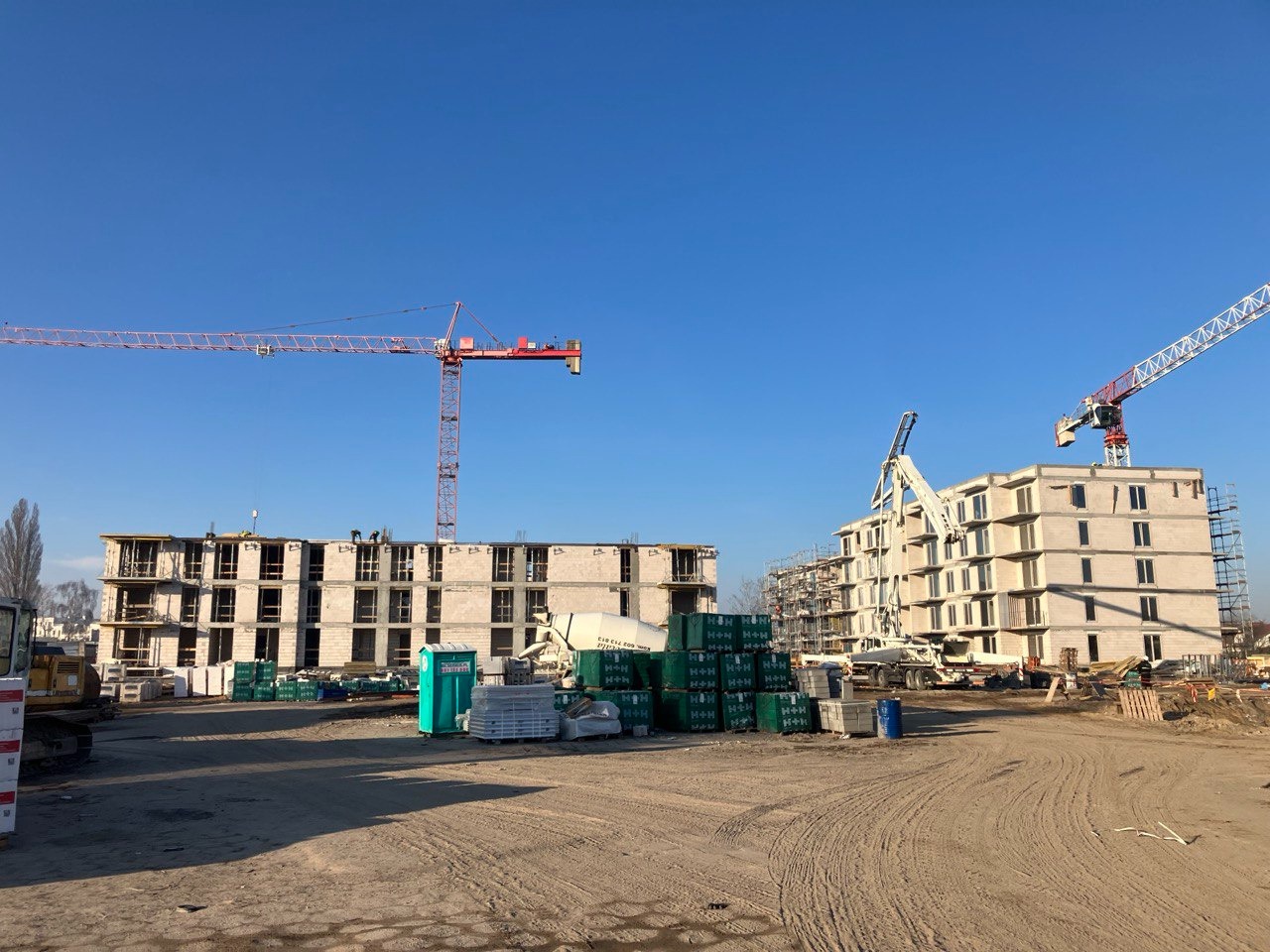 Budowa wielopiętrowego budynku z betonu komórkowego, widoczne rusztowania, dźwigi i materiały budowlane na placu budowy w słoneczny dzień.