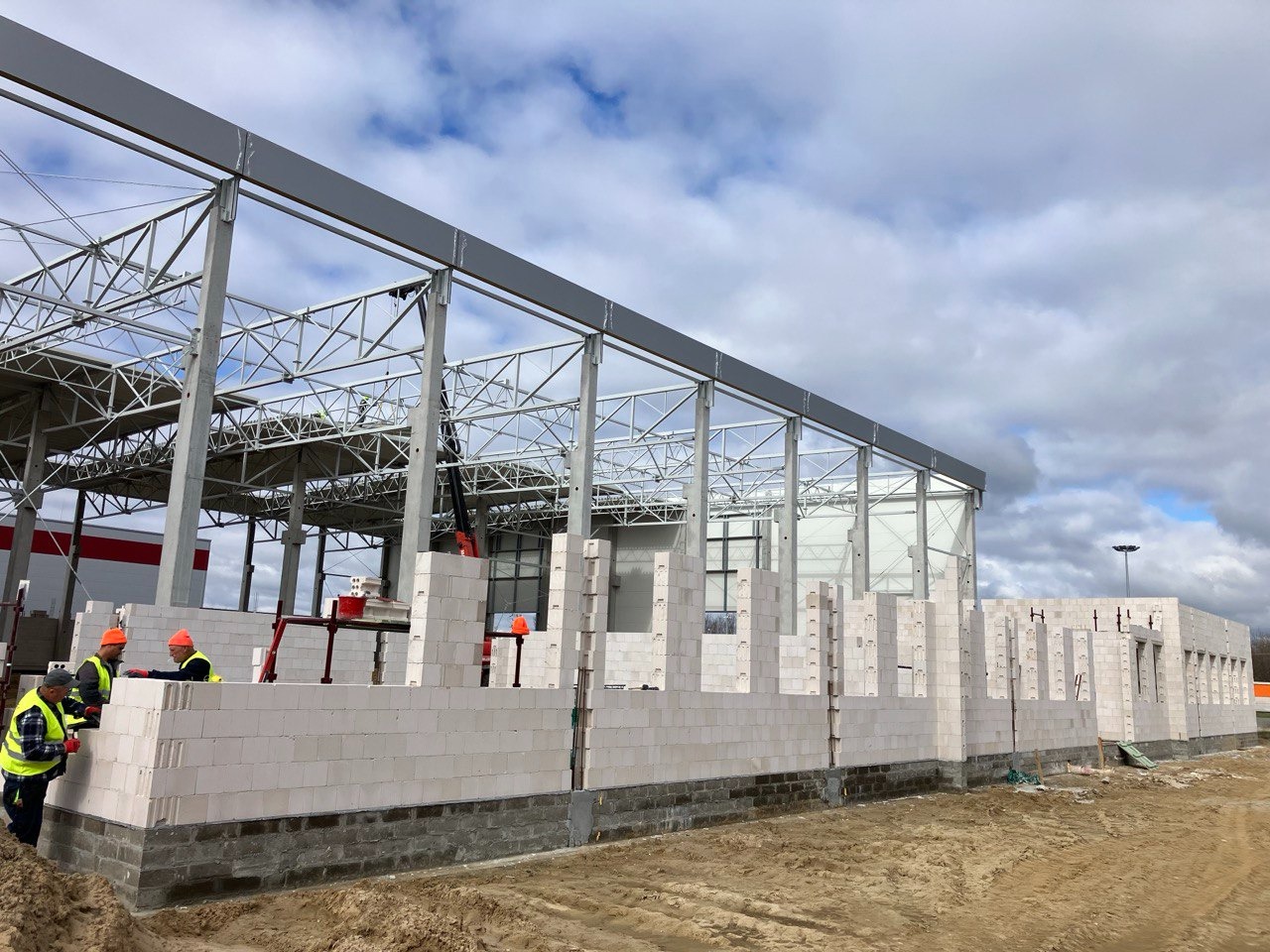 Wznoszenie ścian z białych bloczków na budowie hali przemysłowej z widoczną stalową konstrukcją szkieletową i pracownikami w odblaskowych kamizelkach.