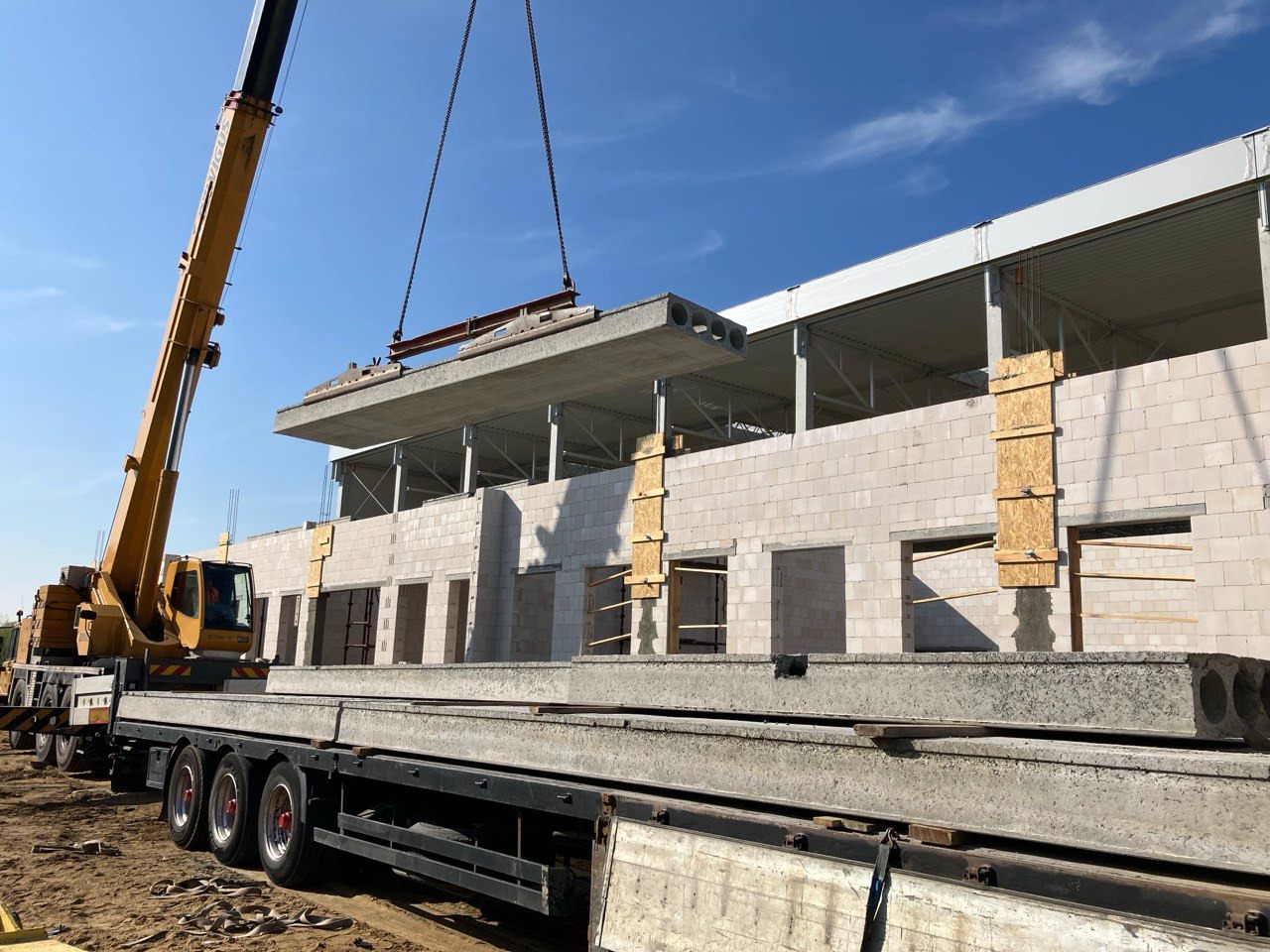 Żuraw przenosi prefabrykowany strop kanałowy na budowę dwukondygnacyjnego budynku z białego bloczka, widoczne szalunki okienne.