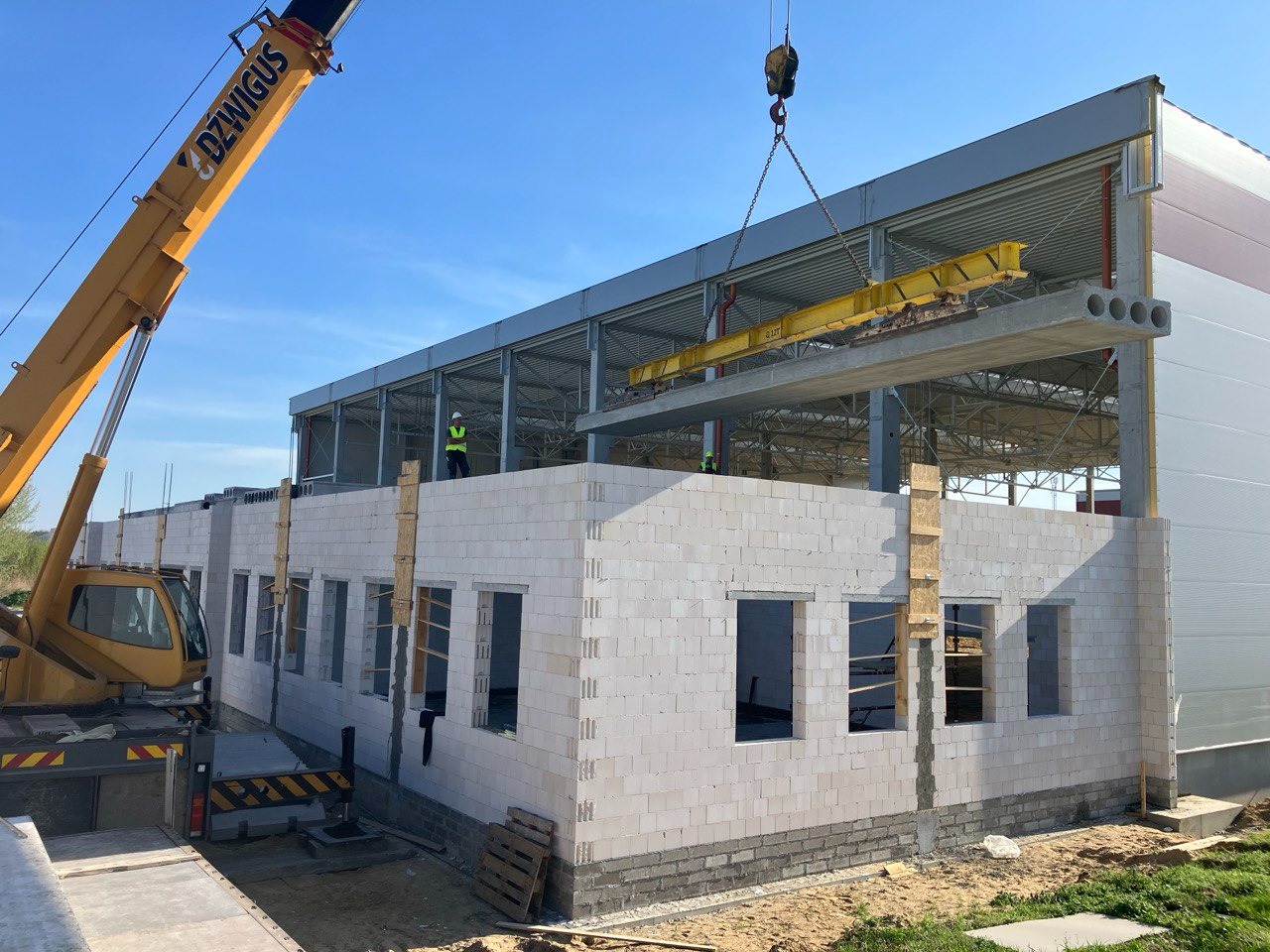 Żuraw przenosi prefabrykowany strop nad budynkiem z białych bloczków, widoczny pracownik w kamizelce odblaskowej, konstrukcja stalowa dachu w tle.