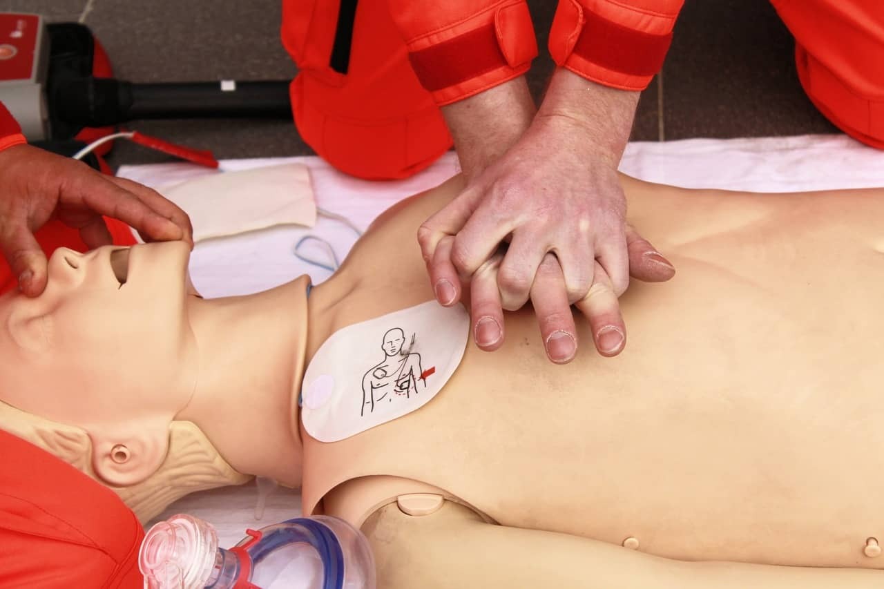 Instruktor demonstruje resuscytację krążeniowo-oddechową na manekinie z naklejonym schematem ułożenia elektrod defibrylatora. Widoczne dłonie, maska i elementy AED.