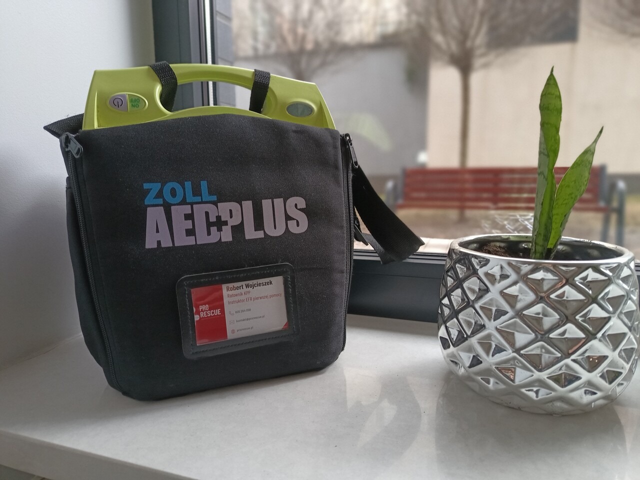 Zestaw AED Plus firmy Zoll w torbie obok rośliny doniczkowej na parapecie. Widoczna etykieta z danymi ratownika KPP i instruktora EFR pierwszej pomocy.