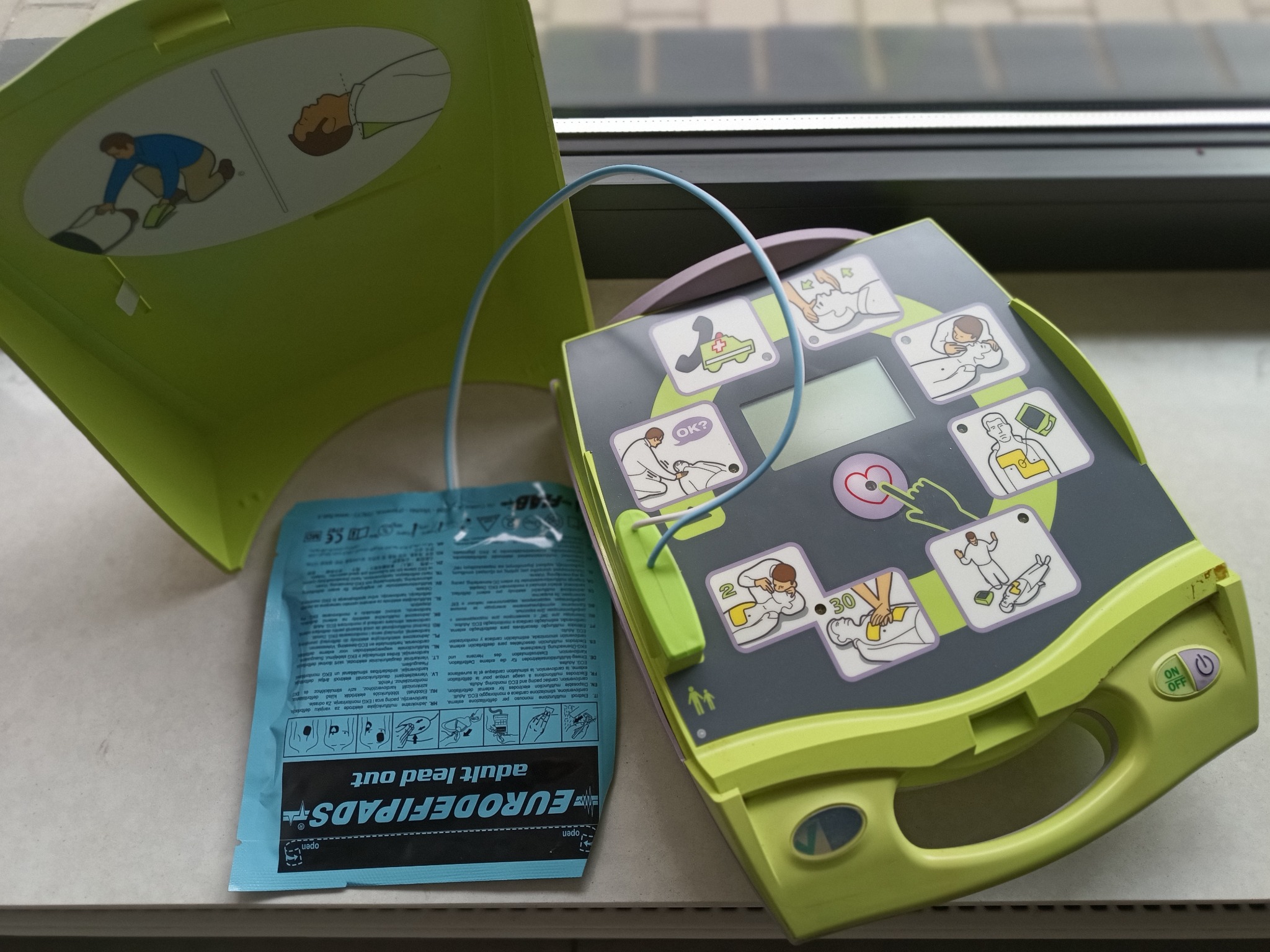 Defibrylator AED z otwartą pokrywą i elektrodami, instrukcja obsługi widoczna na pokrywie, urządzenie na jasnym tle, blisko okna.