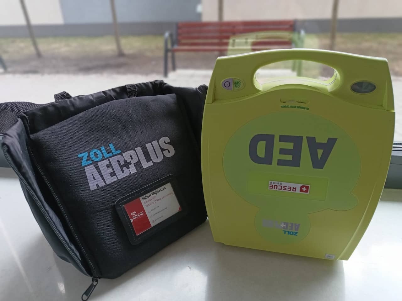 Zestaw AED Zoll: torba z napisem 'ZOLL AECPLUS' i żółty defibrylator z napisem 'AED', na tle okna i ławki. Sprzęt do udzielania pierwszej pomocy.