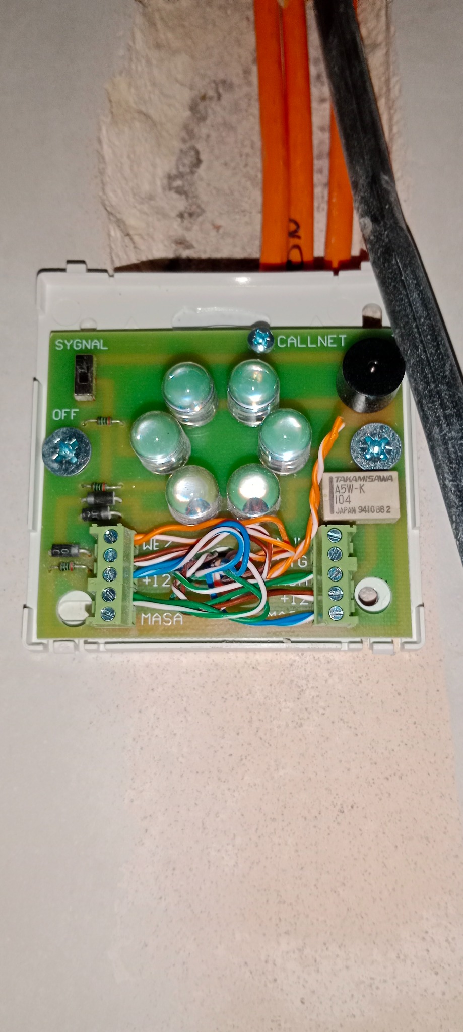 Płyta główna domofonu Callnet z widocznymi elementami elektronicznymi, śrubami, złączami kablowymi i przewodami w kolorach pomarańczowym, niebieskim, białym i zielonym, zamontowana w ścianie...