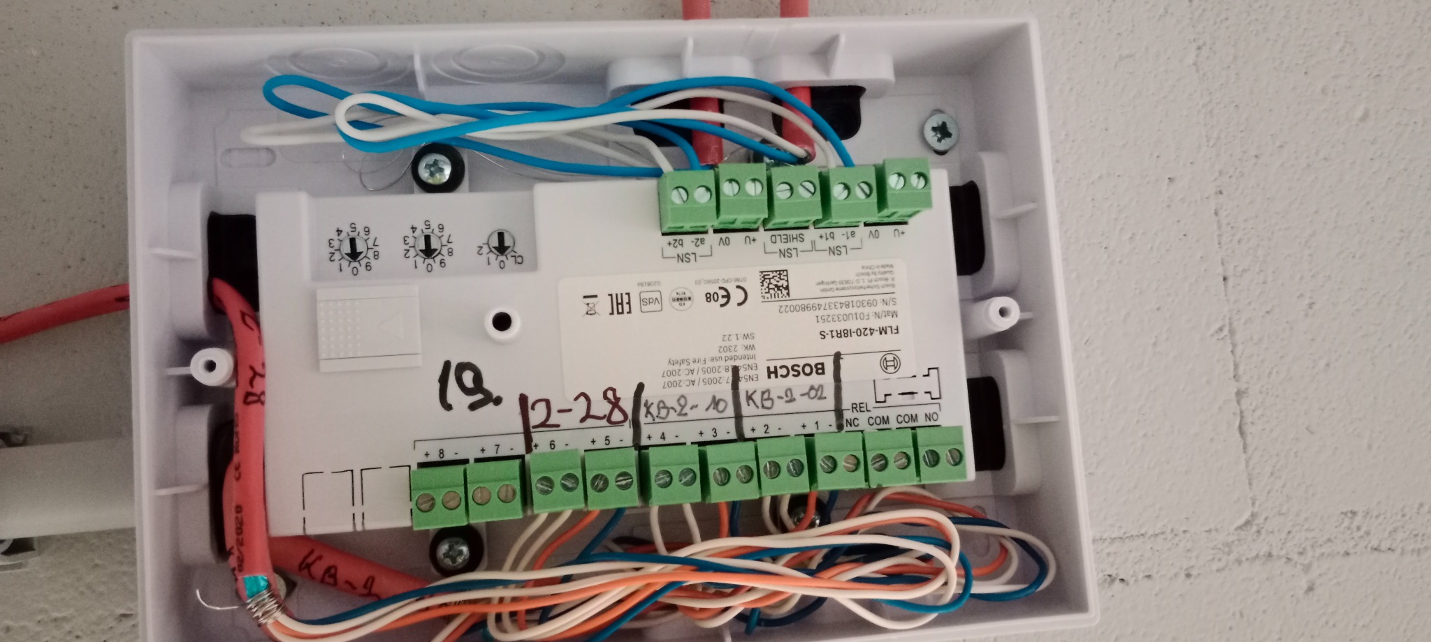 Otwarta, biała puszka instalacyjna Bosch FLM-420-IBR1-S z widocznymi przewodami różnej grubości i kolorów podłączonymi do zielonych złączek, z odręcznym napisem '19. 12-28/KB-2-10 KB-2-02'...