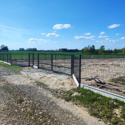 PHU GIFCIK - Nowo postawione, szare ogrodzenie z siatki na betonowej podmurówce, otaczające plac budowy na tle zielonego pola i błękitnego nieba.