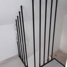 Nowoczesna, czarna balustrada schodowa z pionowych, metalowych pręt&oacute;w, zamontowana w jasnym wnętrzu z drewnianą podłogą.