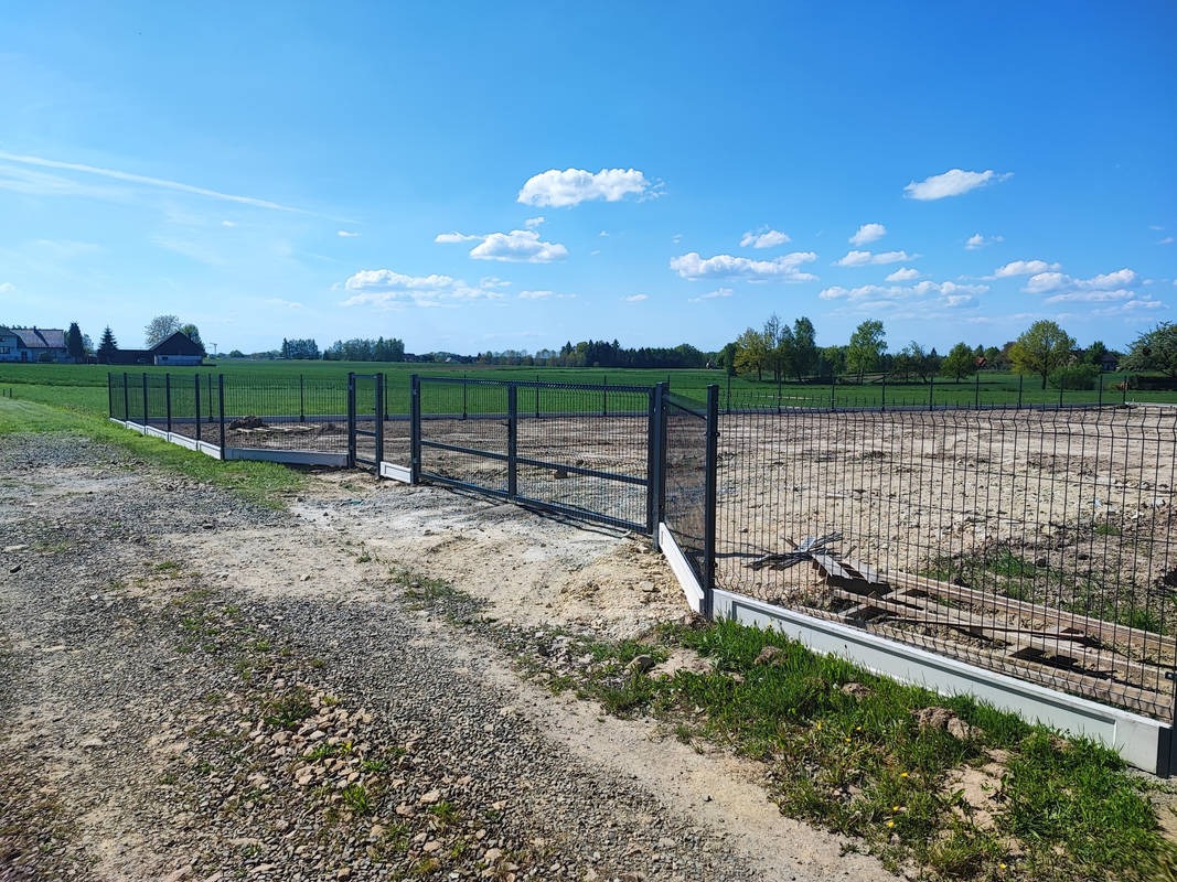 Nowo postawione, szare ogrodzenie z siatki na betonowej podmurówce, otaczające plac budowy na tle zielonego pola i błękitnego nieba.
