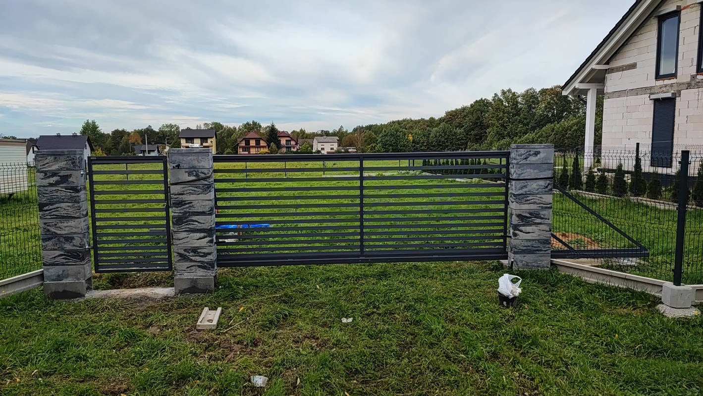Nowoczesna, pozioma brama metalowa z szarymi, modułowymi słupkami z efektem kamienia, częściowo zainstalowana na trawniku przed domem w budowie i ogrodzeniem z siatki.