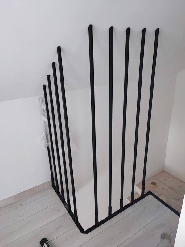 Nowoczesna, czarna balustrada schodowa z pionowych, metalowych prętów, zamontowana w jasnym wnętrzu z drewnianą podłogą.