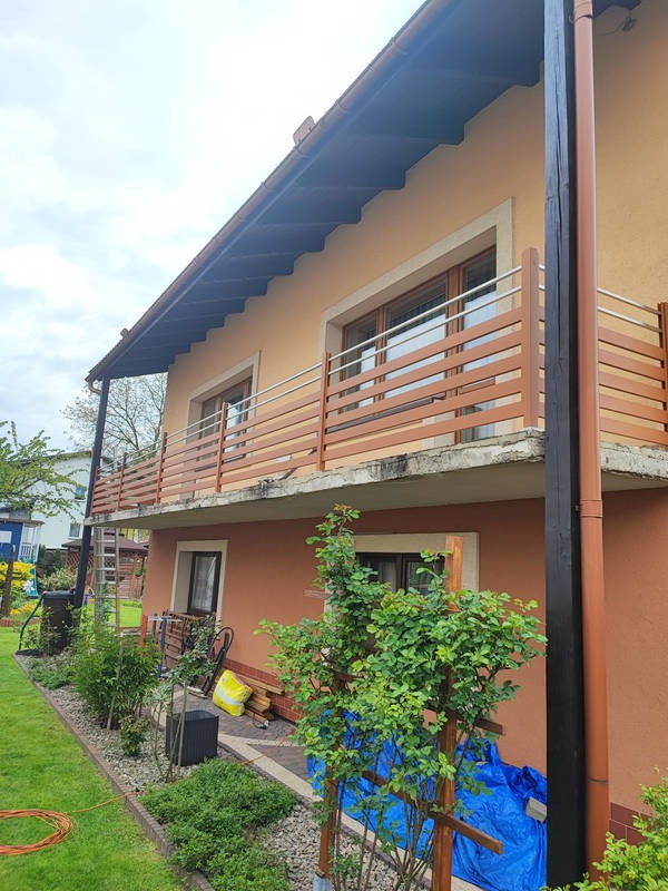 Dwukondygnacyjny dom z balkonami zabezpieczonymi nowoczesnymi balustradami z poziomych, drewnopodobnych listew i metalowych rurek, widok z ogrodu.