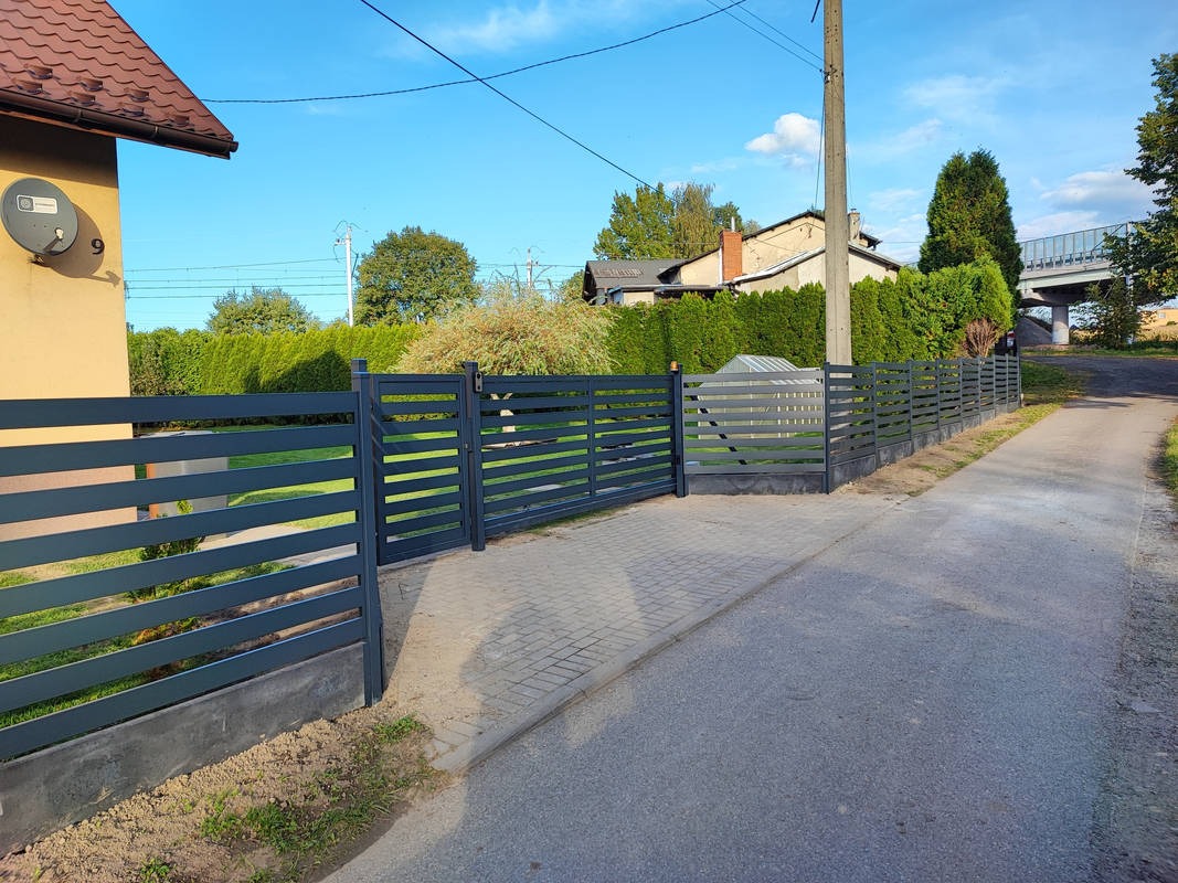 Nowoczesne grafitowe ogrodzenie z poziomych profili, z bramą wjazdową, na tle zadbanego ogrodu i domu z anteną satelitarną.