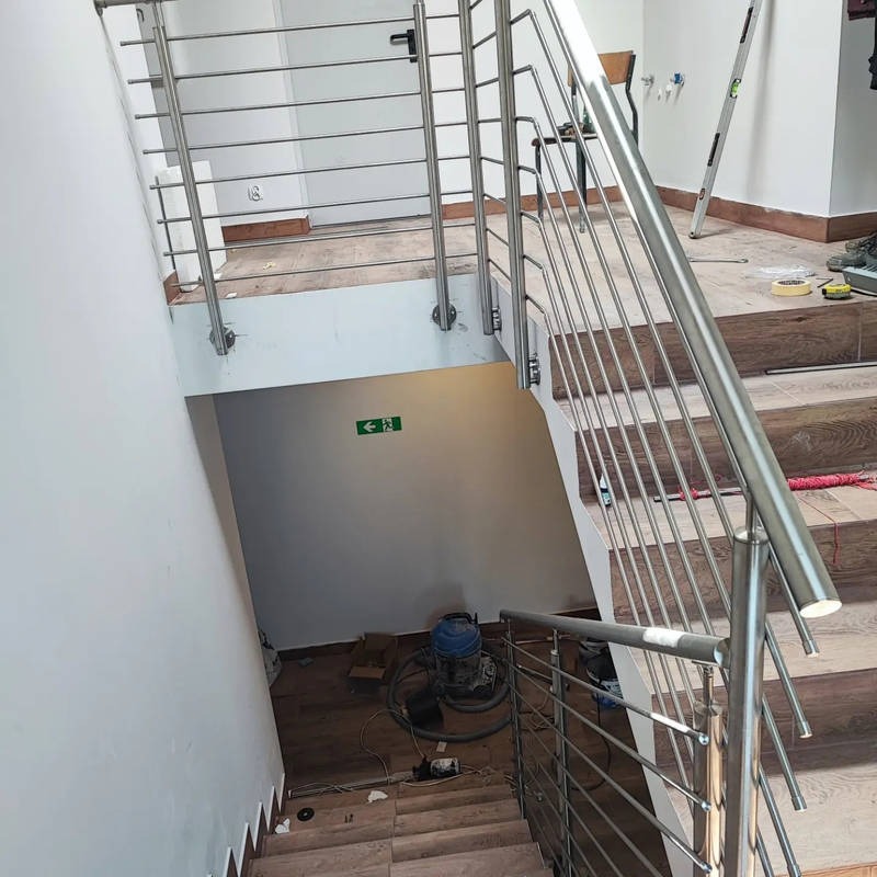 Wnętrze w trakcie wykańczania: schody z drewnopodobnymi płytkami i balustradą ze stali nierdzewnej, widoczny sprzęt budowlany i znak ewakuacyjny.