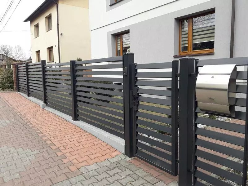 Nowoczesne ogrodzenie panelowe z poziomych, szarych profili, brama z metalową skrzynką na listy, na tle domu z szarą elewacją.
