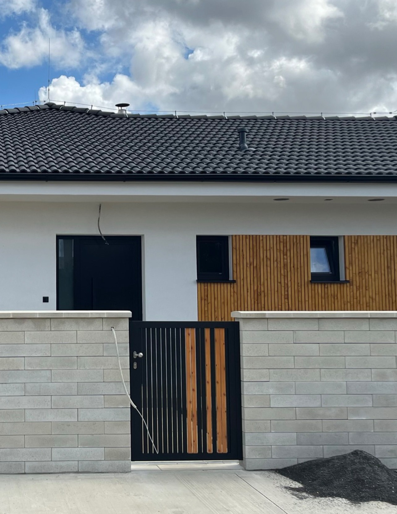 Nowoczesna brama z czarnymi, pionowymi prętami i drewnianymi akcentami, wbudowana w ogrodzenie z szarej, betonowej kostki, na tle białego domu z drewnianą elewacją i czarnymi oknami.