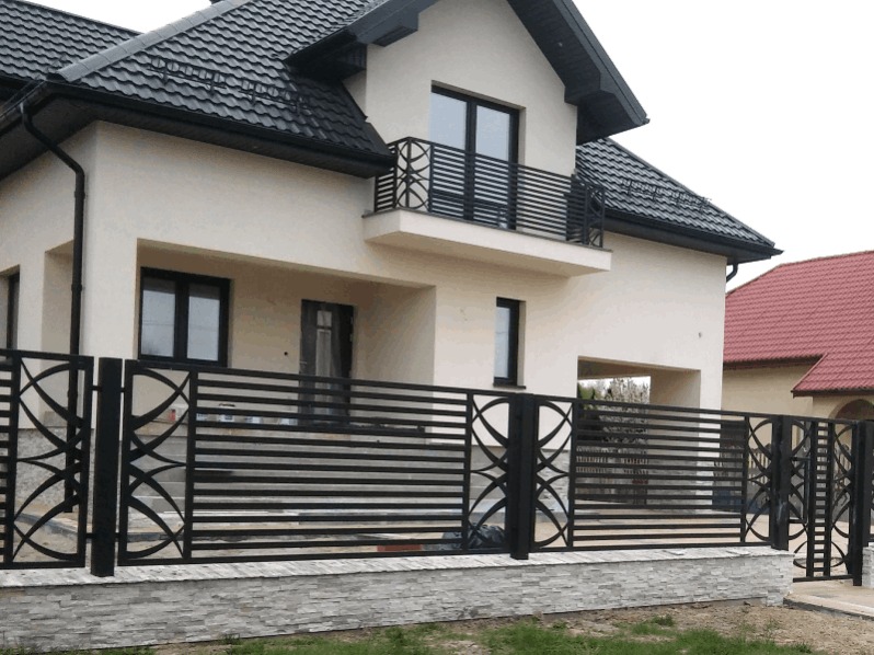 Nowoczesne czarne ogrodzenie metalowe z poziomymi prętami i dekoracyjnymi elementami w kształcie okręgów, zamontowane na niskim murku z szarej imitacji kamienia, na tle dwukondygnacyjnego domu...