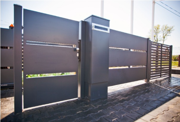 Nowoczesna, szara brama wjazdowa z poziomymi panelami i skrzynką na listy z numerem domu, uzupełniona o przylegające ogrodzenie lamelowe, na tle kostki brukowej.