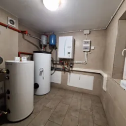 Kotłownia z dwoma białymi zbiornikami, niebieskimi naczyniami przeponowymi, kotłem gazowym, licznikiem i instalacją hydrauliczną w pomieszczeniu wyłożonym kafelkami.