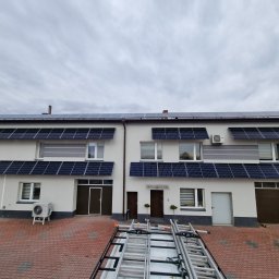 Solarid - Panele fotowoltaiczne zamontowane nad oknami i na dachu segmentu domu jednorodzinnego, na pierwszym planie drabiny na przyczepce samochodowej, dodatkowo widoczny klimatyzator na zewnątrz budynku.