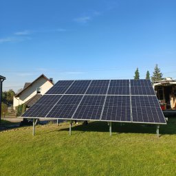 Solarid - Panel fotowoltaiczny ustawiony na trawniku przed domem jednorodzinnym, widoczny fragment samochodu dostawczego w tle, błękitne niebo.