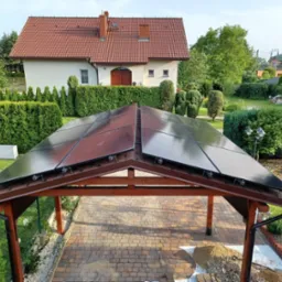 Panel słoneczny zamontowany na drewnianej wiacie garażowej, widok z góry na kostkę brukową, w tle dom jednorodzinny z czerwonym dachem.