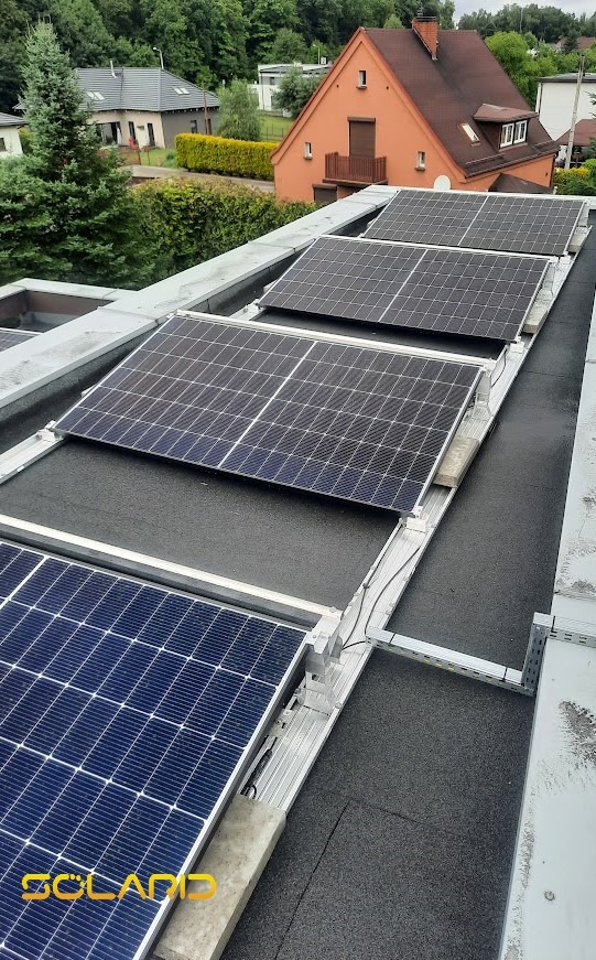 Panele fotowoltaiczne na płaskim dachu z widoczną konstrukcją montażową i fragmentem logo firmy SolarD w lewym dolnym rogu, w tle domy jednorodzinne.